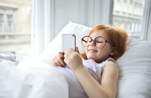 使用智能手机时躺在床上的白色背心的微笑女孩的照片 · 免费素材图片