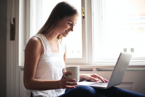 微笑的女人在白色上衣，坐在窗户旁边，同时使用一台笔记本电脑和拿着一个白色的陶瓷杯的侧视图照片 · 免费素材图片