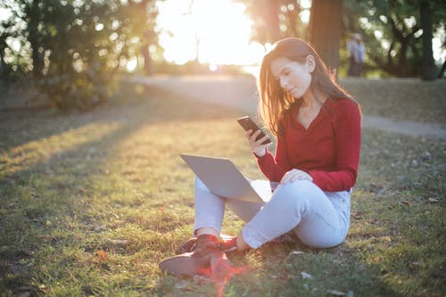 女人穿着一件红色的毛衣和蓝色的裤子，坐在绿草地上发短信，同时在她的膝盖上携带一台笔记本电脑的选择性焦点照片 · 免费素材图片