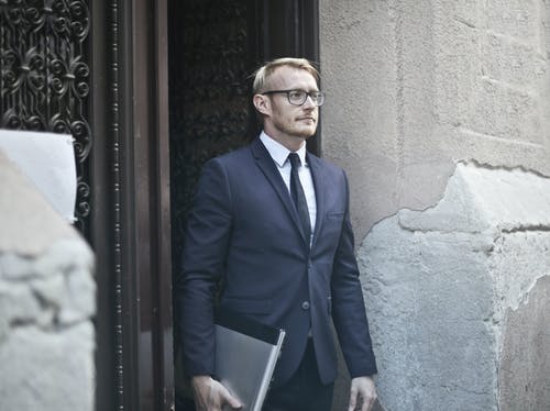 蓝色西装外套和眼镜站在门口，拿着一台笔记本电脑的人的照片 · 免费素材图片
