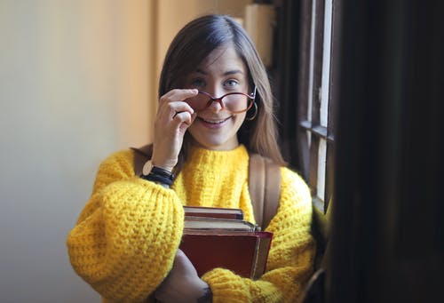 微笑的女人，穿着黄色的毛衣和黑色镜框的眼镜，拿着书时站在窗户旁边的肖像照片 · 免费素材图片
