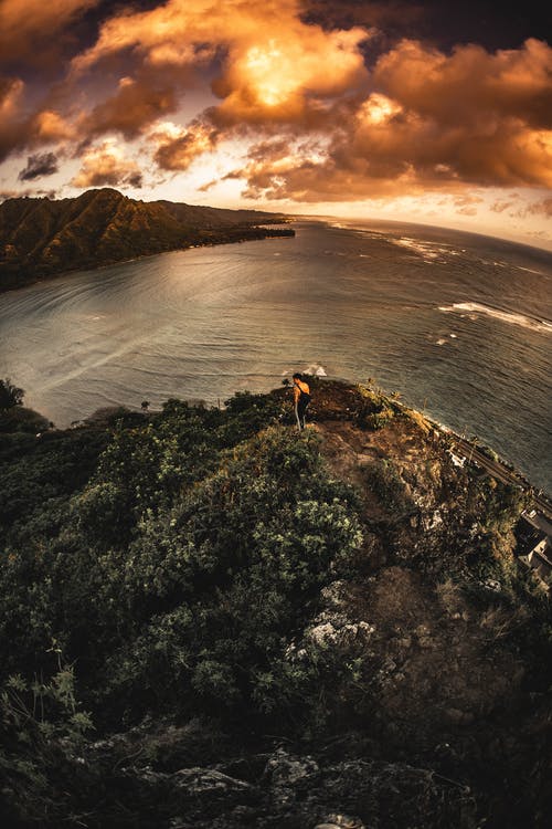 匿名的徒步旅行者在日落期间在风景如画的海中达到岩石峭壁的顶峰 · 免费素材图片
