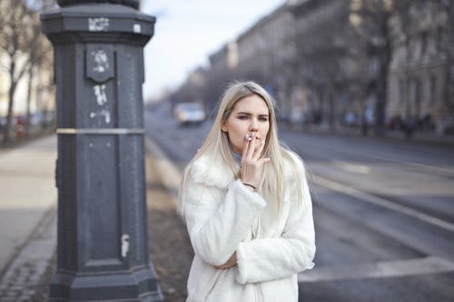 女人穿着白色外套站在路边抽着烟的选择性焦点照片 · 免费素材图片