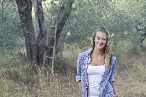 蓝色衬衫和白色上衣站在橄榄树林中的微笑女人的照片 · 免费素材图片