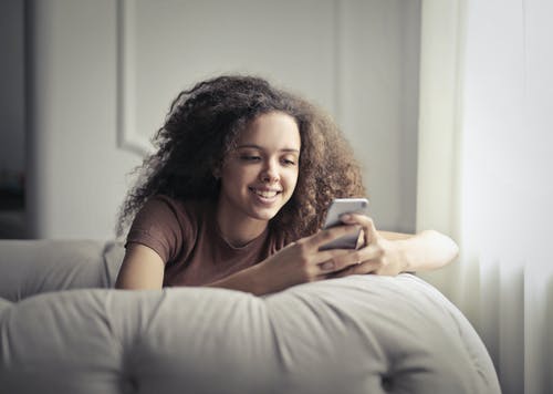 微笑的女人坐在灰色的豆袋上，同时使用她的手机的照片 · 免费素材图片