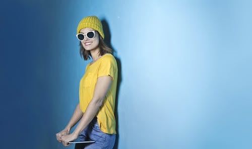 微笑的女人，穿着黄色的t恤和无檐小便帽，蓝色的牛仔裤和白色太阳镜站在平板电脑时站在蓝墙前的照片 · 免费素材图片