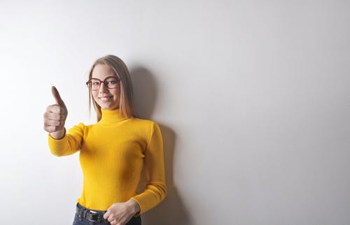 黄色高领毛衣，蓝色牛仔牛仔裤和眼镜放弃拇指的女人的照片 · 免费素材图片