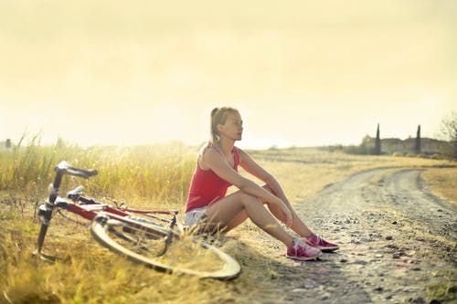 女人穿着红色背心坐在她的自行车旁边的土路边的照片 · 免费素材图片