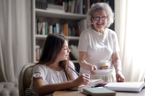 给曲奇饼和杯热的饮料的愉快的年迈的妇女在家做家庭作业的喜悦的孙女 · 免费素材图片