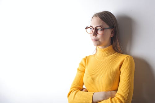 女人穿着黄色的高领毛衣和黑色镜框的眼镜，看着别处时站在白墙前的肖像照片 · 免费素材图片