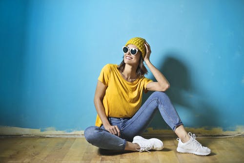 微笑的女人，穿着黄色的t恤和无檐小便帽帽子，蓝色的牛仔裤和白色的太阳镜，坐在蓝色的墙前的地板上的照片 · 免费素材图片