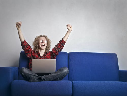 激动的人，举起手来坐在蓝色的沙发上，同时使用一台笔记本电脑的照片 · 免费素材图片