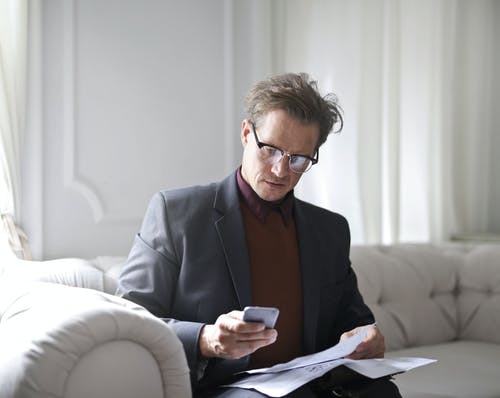 穿着西装和黑色镜框的眼镜在使用电话时坐在白色沙发上的男人的照片 · 免费素材图片
