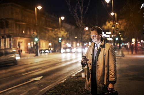 穿着棕色大衣的人拿着公文包，晚上站在人行道上时使用他的电话的照片 · 免费素材图片