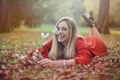 笑的女人，穿着红色的长袖衣服，躺在干燥的叶子，同时拿着一根棍子与干树叶笑的选择性焦点照片 · 免费素材图片