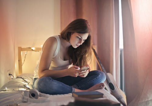 女人穿着白色背心和蓝色牛仔牛仔裤，坐在床上，一边用她的手机的照片 · 免费素材图片