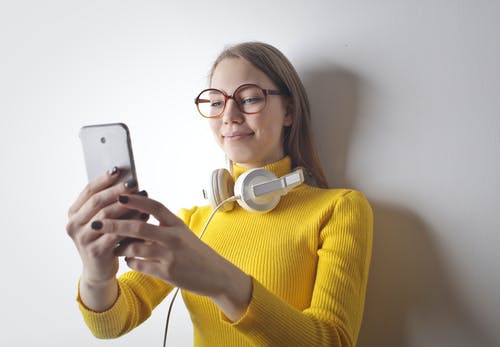 黄色高领毛衣的微笑的女人，用她的手机在她脖子上的耳机的照片 · 免费素材图片