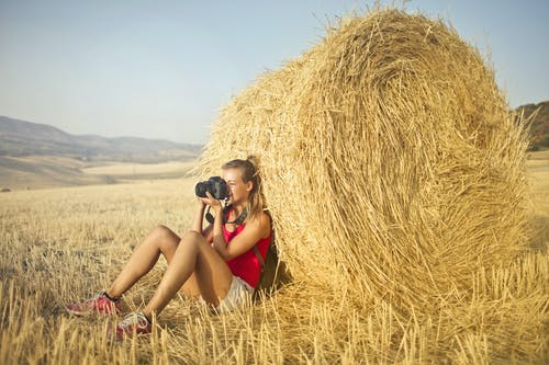 穿红色背心和白色短裤的女人坐在地上，一边拍照一边倾斜一堆干草 · 免费素材图片
