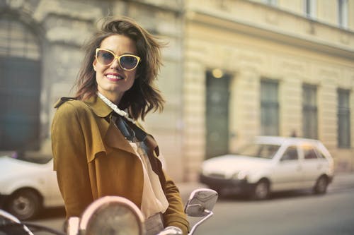 微笑的女人，穿着棕色外套和黄色太阳镜站在人行道上的选择性焦点照片 · 免费素材图片
