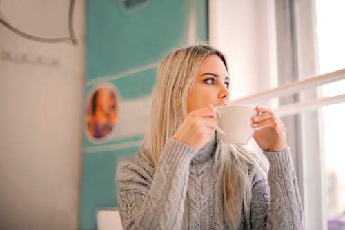 选择性焦点的灰色毛衣，看着别处时从白色陶瓷杯喝水的女人的照片 · 免费素材图片