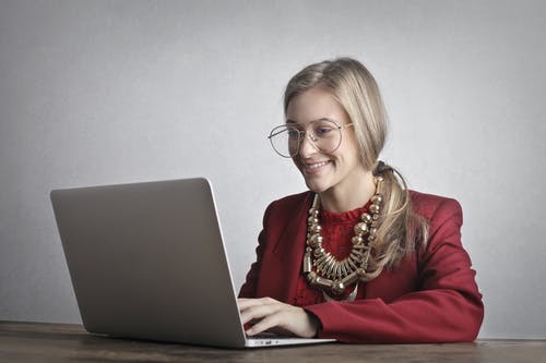微笑的女人，穿着红色外套和眼镜使用一台笔记本电脑的照片 · 免费素材图片