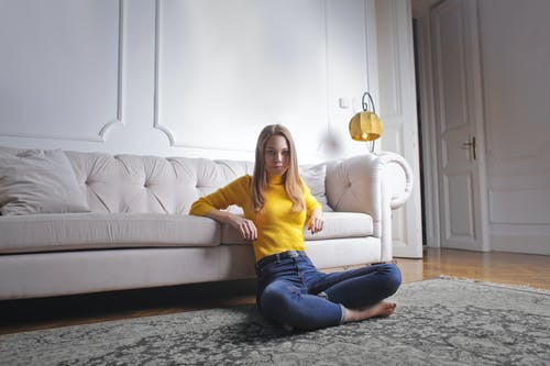 穿着黄色高领毛衣和蓝色牛仔牛仔裤的女人坐在地板上，双腿交叉而斜倚在白色沙发上的照片 · 免费素材图片