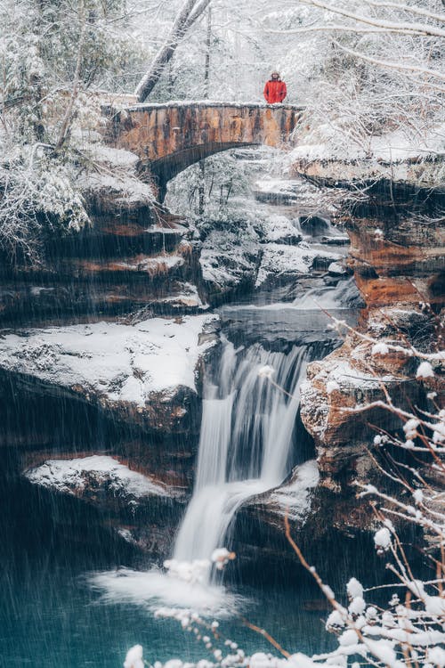 站在水瀑布附近的桥上的红色外套的人 · 免费素材图片