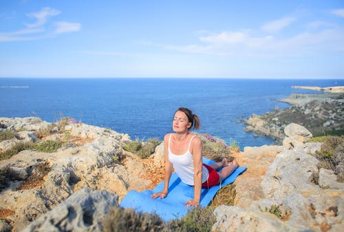 白色背心在岩石海岸上做瑜伽运动的女人 · 免费素材图片