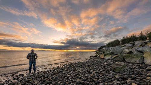 日落期间站在多岩石的海岸上的人 · 免费素材图片