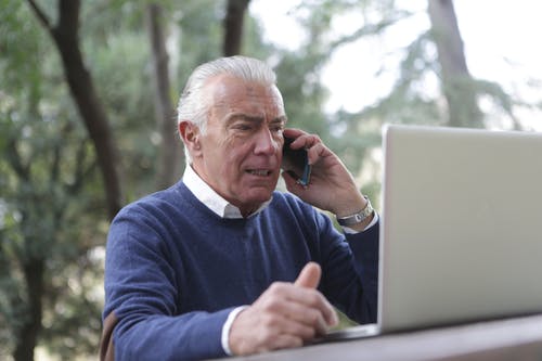 坐在桌子旁使用笔记本电脑和手机的蓝色毛衣的男人 · 免费素材图片