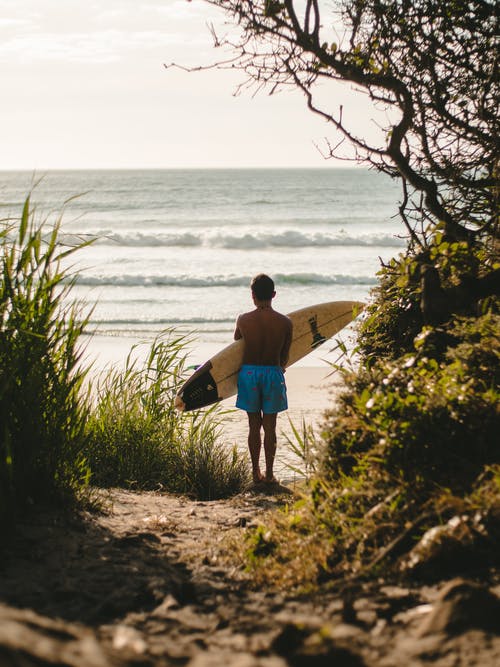蓝色的短裤，拿着棕色的冲浪板，在海边散步的人 · 免费素材图片