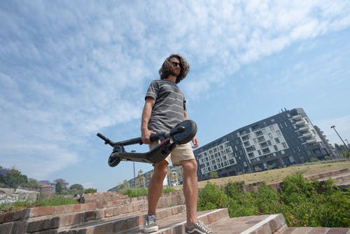 灰色t恤和短裤的男人抱着折叠电动滑板车 · 免费素材图片