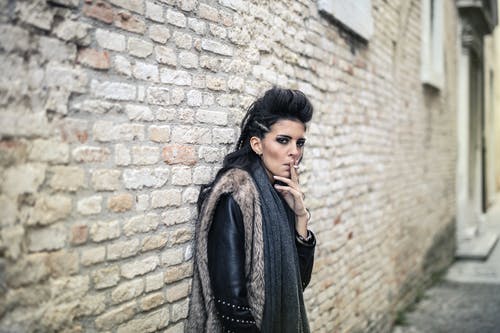 在砖墙附近吸烟时穿皮草大衣的女人的照片 · 免费素材图片