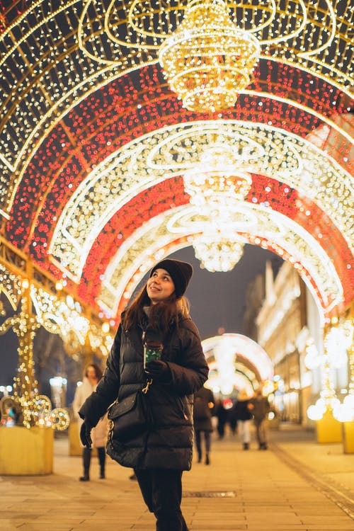 黑夹克的女人站在反对点燃的圣诞灯拱 · 免费素材图片