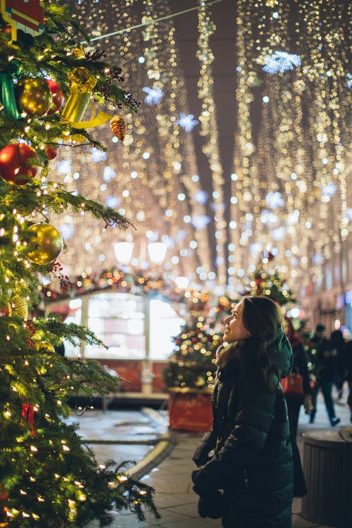 站在绿色的圣诞树，与串灯旁边的黑色外套的女人 · 免费素材图片