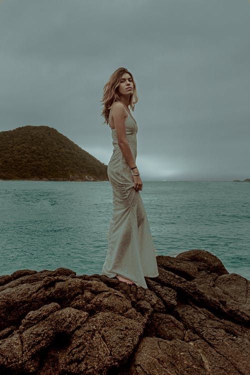 白色无袖连衣裙站在水体附近的棕色岩石上的女人 · 免费素材图片