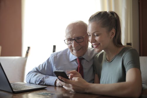 年轻积极的女士坐在笔记本电脑上向老人显示在智能手机上的照片 · 免费素材图片