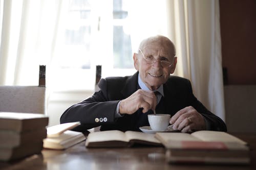 正式衣服的老人喝热的饮料，当坐在与书的桌上时 · 免费素材图片