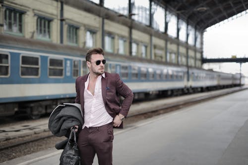 站立在有外套和旅行袋子的铁路平台上的确信的时髦男性旅客 · 免费素材图片