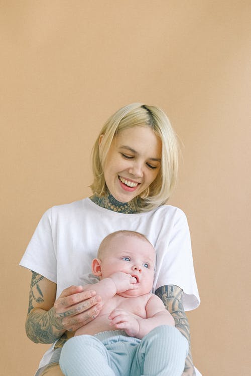 拥抱逗人喜爱的新生儿的笑的年轻母亲，当握在嘴的婴孩手指在托儿所时 · 免费素材图片