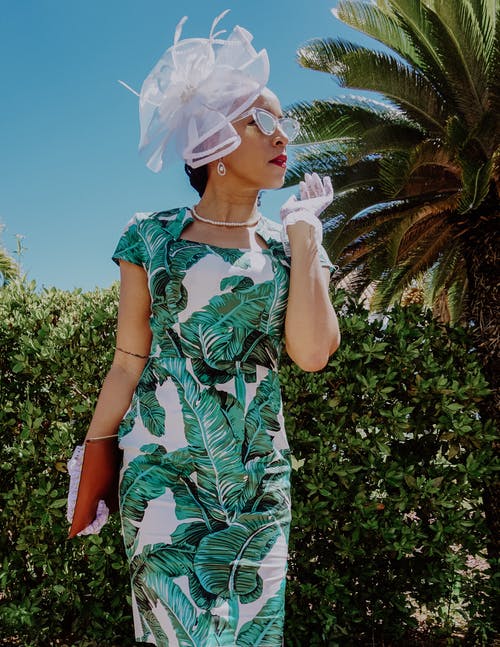 绿色和白色的花裙子，戴着白色口罩站在绿色的棕榈树附近的女人 · 免费素材图片