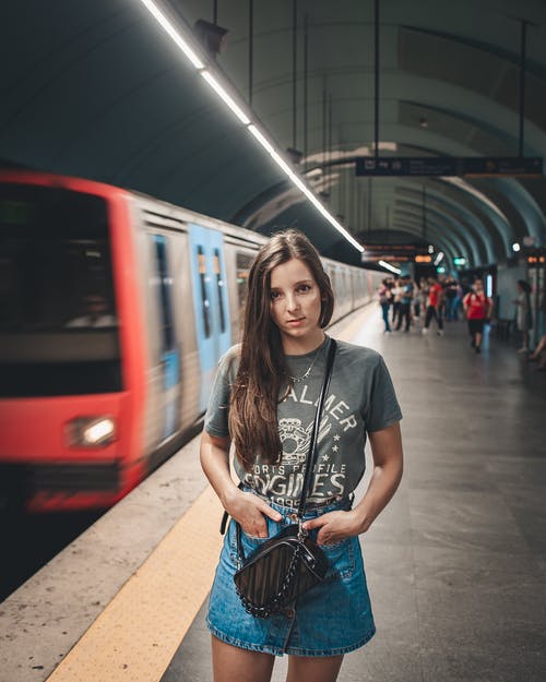 身穿灰色圆领t恤和蓝色牛仔裙的女人站在地铁系统中的火车附近 · 免费素材图片