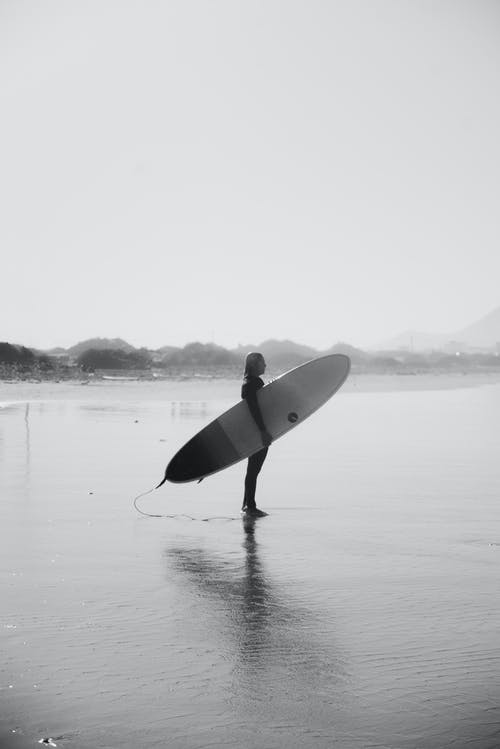 男子手持冲浪板站在海滩上的灰度照片 · 免费素材图片