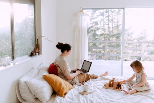 可爱的蹒跚学步的女孩玩木积木坐在床上，而母亲在阳光明媚的早晨使用笔记本电脑 · 免费素材图片