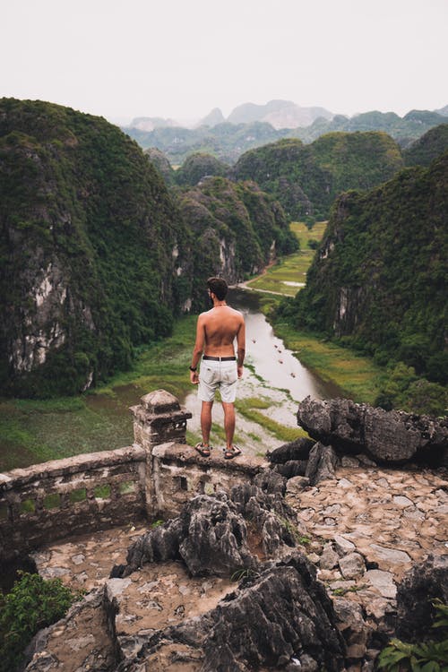 白色短裤的男人站在绿山附近的棕色岩石上 · 免费素材图片