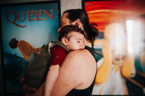 族裔父亲背着熟睡的婴儿与海报墙附近 · 免费素材图片