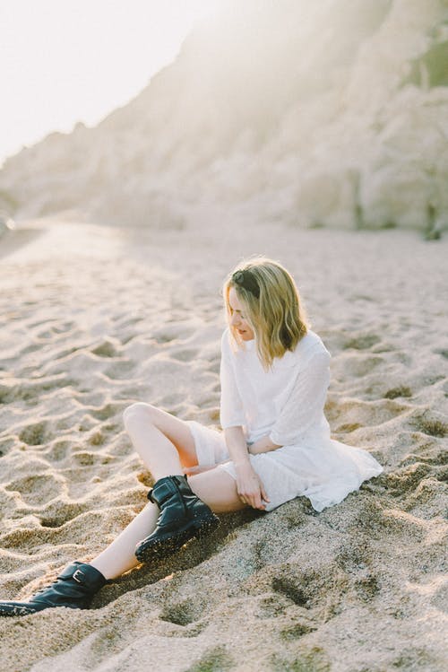 白色长袖衬衫和蓝色牛仔牛仔裤，坐在灰色的沙滩上的女人 · 免费素材图片