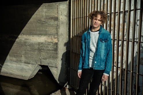 站在灰色铁门旁边的蓝色牛仔夹克的男人 · 免费素材图片