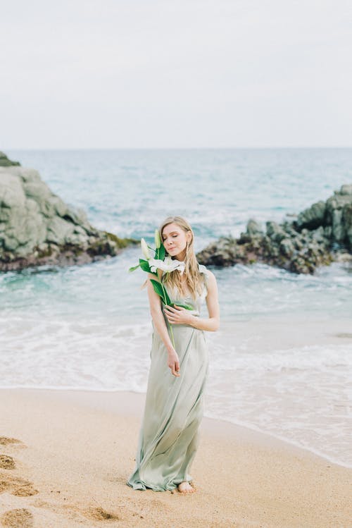 米色的连衣裙，拿着花束站在海滩上的女人 · 免费素材图片