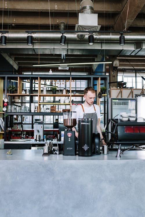 穿着灰色圆领t恤的男人站在黑色和银色咖啡机附近 · 免费素材图片